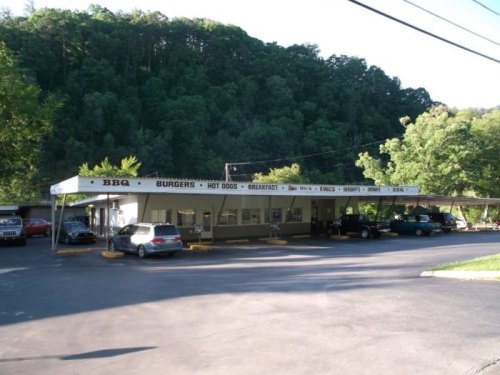 A roadside BBQ restaurant with a covered patio, surrounded by trees and parked cars in a rural setting.