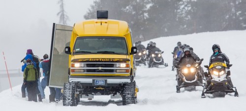 A yellow snow coach parked on a snowy path, with snowmobiles and people in winter gear nearby.
