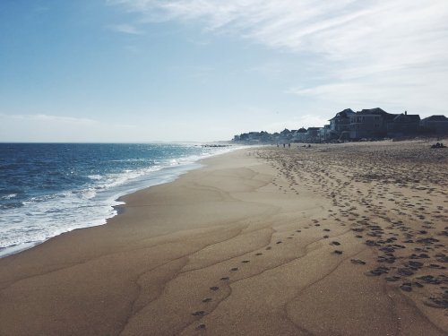 A sandy beach with gentle waves, footprints in the sand, and houses in the distance under a clear blue sky.