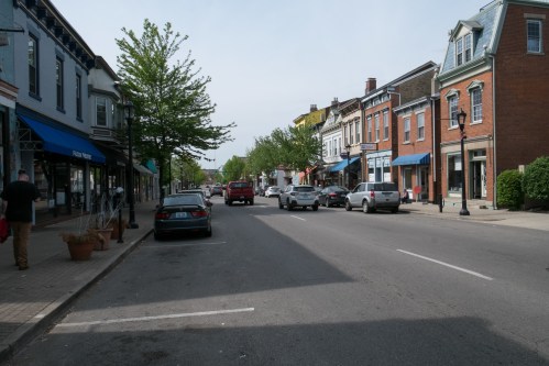 A quiet street lined with shops and trees, featuring parked cars and a few pedestrians on a sunny day.