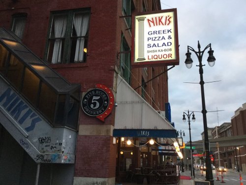 A restaurant exterior featuring a sign for "Niki's Greek Pizza & Salad" and a nearby "Five Bucks Brewery" sign.