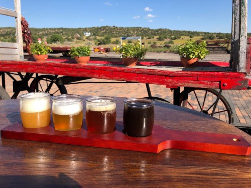 A wooden flight board with four small glasses of beer, set outdoors with potted plants and a scenic background.