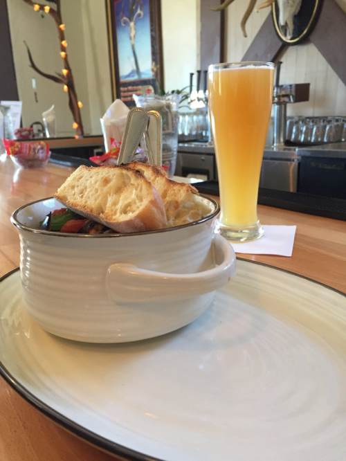 A bowl of stew with bread on top, next to a tall glass of yellow beverage, set on a wooden bar counter.