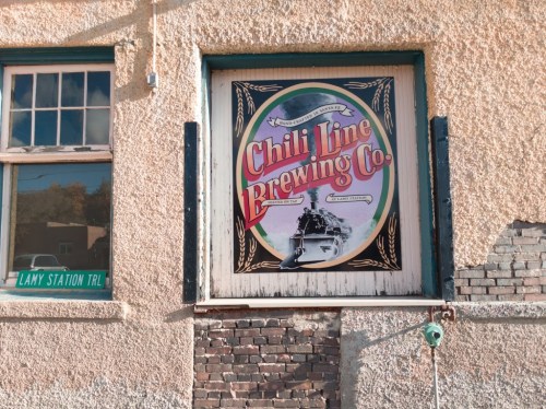 A colorful sign for Chili Line Brewing Co. on a textured wall, with a vintage train image and nearby Lamy Station Trail sign.