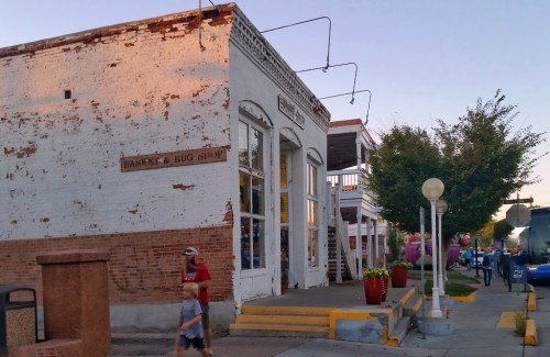 A quaint basket and rug shop with a weathered exterior, surrounded by trees and a sidewalk, during sunset.