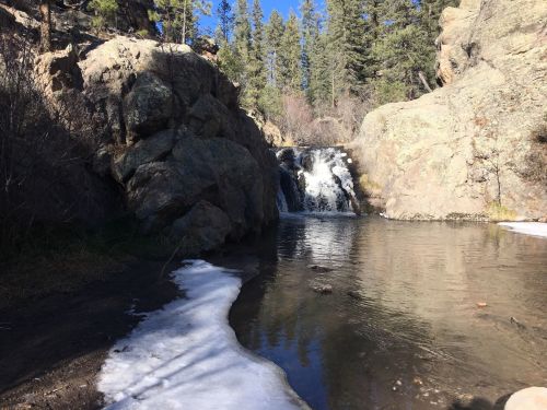 A serene waterfall cascades into a calm pool, surrounded by rocky terrain and evergreen trees under a clear blue sky.