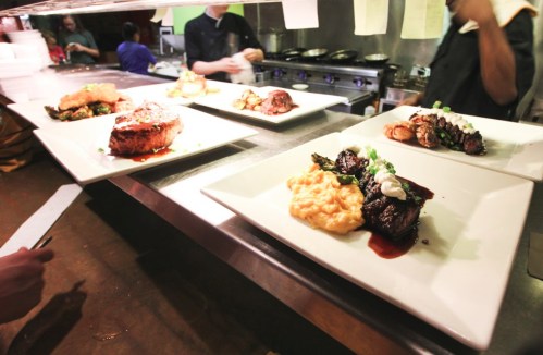 Plated gourmet dishes on a kitchen counter, featuring meats, sides, and garnishes, with chefs in the background.
