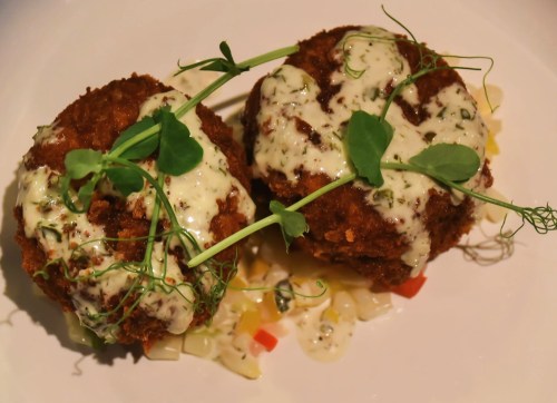 Two golden-brown fried balls topped with creamy sauce and microgreens, served on a colorful vegetable bed.