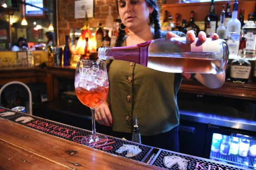 A bartender pours sparkling wine into a glass filled with ice, surrounded by various bottles on a bar.