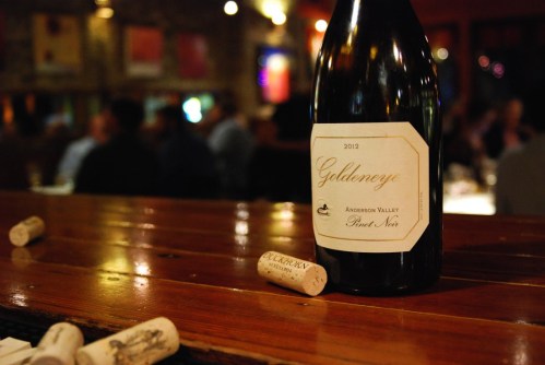 A bottle of 2012 Goldeneye Pinot Noir on a wooden table, surrounded by wine corks in a dimly lit restaurant.