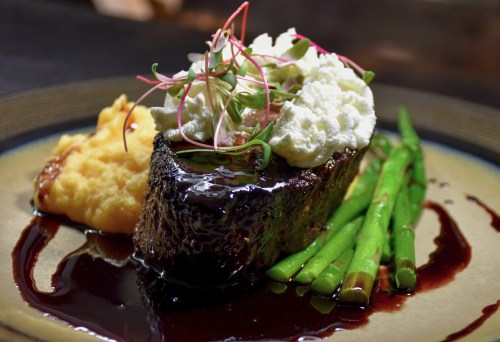 A gourmet plate featuring steak with a rich sauce, mashed potatoes, green beans, and garnished with microgreens.