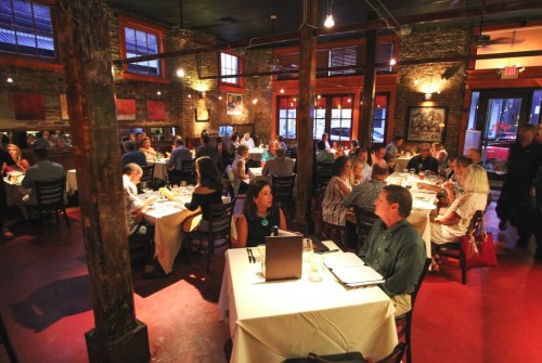A bustling restaurant interior with diners at tables, warm lighting, and exposed brick walls creating a cozy atmosphere.