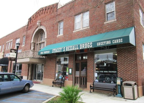 A brick building with a green awning reading "Stacey's Rexall Drugs" and a vintage soda fountain in the window.