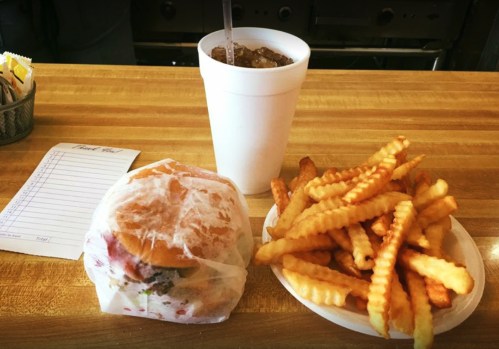 A wrapped burger sits next to a plate of crinkle-cut fries and a large drink on a wooden table.