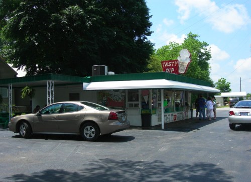 A small ice cream stand with a green roof, featuring a sign that says "Tasty Dip," and a car parked nearby.