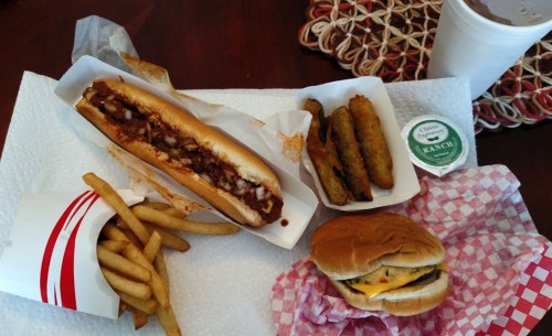 A meal featuring a hot dog with toppings, fries, fried pickles, and a cheeseburger on a table with a drink.