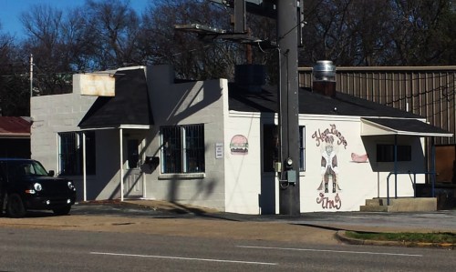A small, white building with a mural, featuring a burger and a cowboy, located on a street corner.