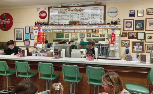 A retro diner interior with a long counter, green stools, and staff serving customers. Walls adorned with memorabilia.