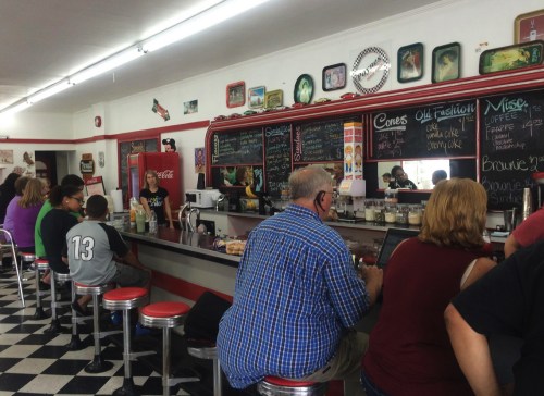 A retro diner with red accents, featuring patrons at the counter, a chalkboard menu, and a checkered floor.