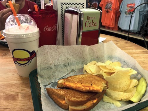 A tray with a grilled cheese sandwich, chips, pickles, a milkshake, and a red drink in a diner setting.