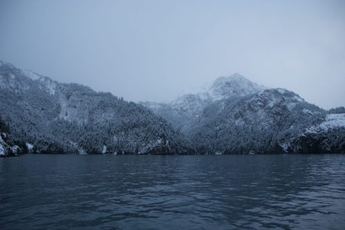 A serene winter landscape featuring snow-covered mountains reflected in calm water under a gray sky.