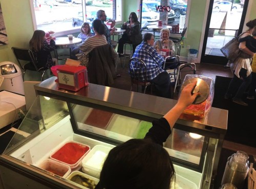 A person serves ice cream at a shop, with customers seated at tables in the background enjoying their treats.