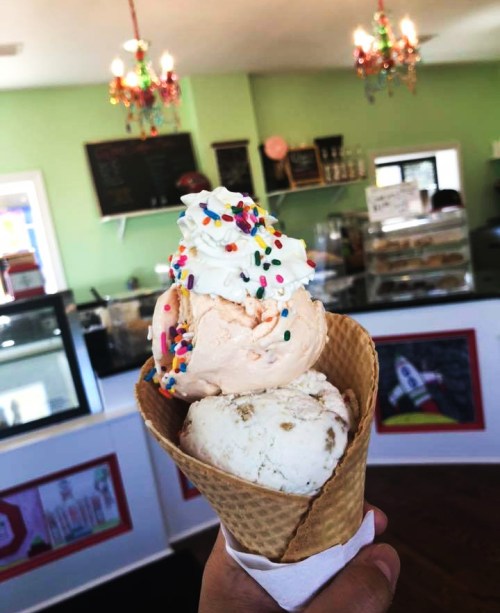 A hand holds a two-scoop ice cream cone with sprinkles and whipped cream, set against a colorful ice cream shop interior.