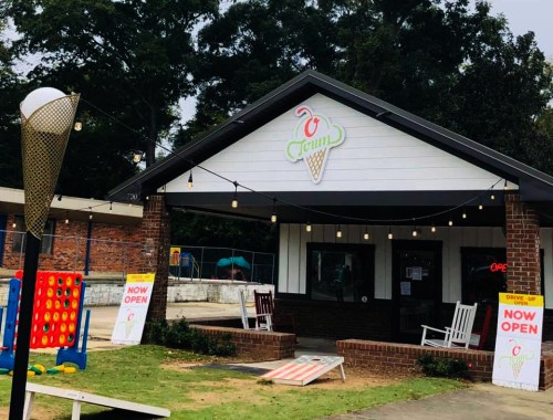 A charming ice cream shop with a sign reading "Now Open," featuring outdoor seating and a colorful game area.