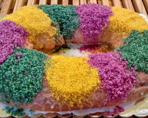 A colorful king cake topped with purple, green, and yellow sugar, sitting on a wooden surface.