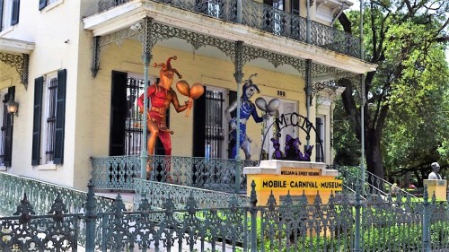 Colorful jester murals on a historic building, featuring a sign for the Mobile Carnival Museum. Lush greenery surrounds it.