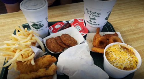 A tray of fast food featuring fries, chicken tenders, onion rings, dipping sauces, and drinks.