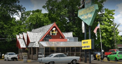 A retro-style diner with a peaked roof, large windows, and a sign featuring a cartoon chef, surrounded by trees and cars.