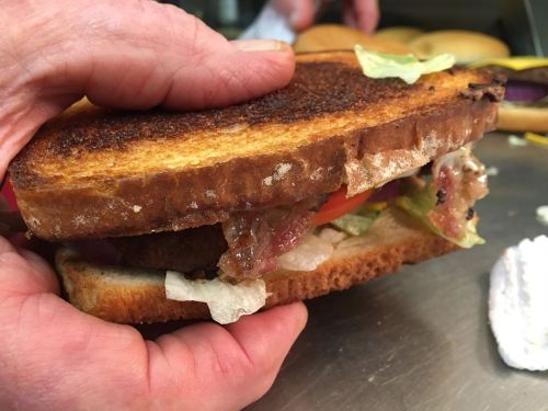 A hand holding a toasted sandwich filled with lettuce, tomato, and bacon, with other sandwiches in the background.