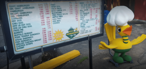 Menu board featuring various food items and prices, with a cartoon duck wearing a chef's hat beside it.