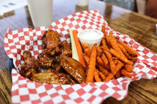 A plate of chicken wings with sauce, sweet potato fries, celery sticks, and a small cup of ranch dressing on a checkered paper.