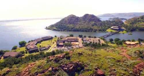 Aerial view of a lakeside resort surrounded by hills and greenery, with buildings and a bridge visible.