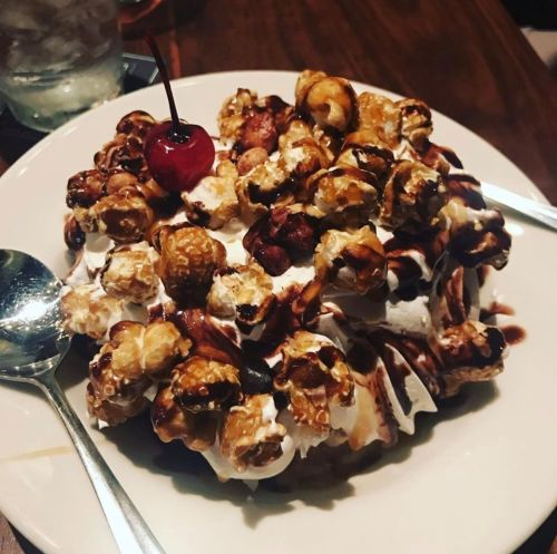 A dessert featuring a mound of popcorn topped with whipped cream, chocolate sauce, and a cherry on a white plate.