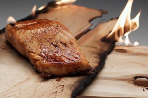 A piece of grilled salmon on a charred wooden plank, with flames in the background.