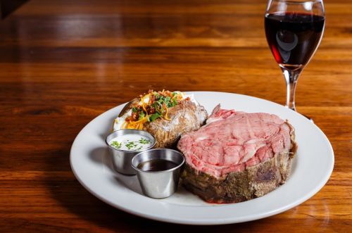 A plate with sliced prime rib, a baked potato topped with sour cream and chives, and a glass of red wine.