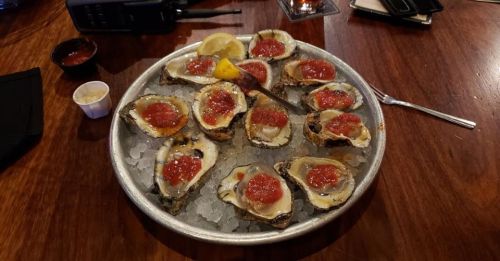 A platter of oysters on ice, topped with cocktail sauce and a lemon wedge, served on a wooden table.