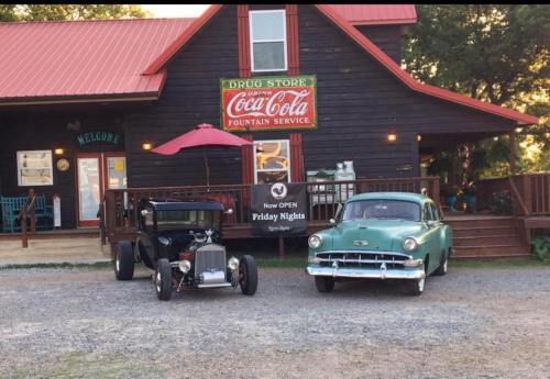 Two vintage cars parked in front of a rustic building with a Coca-Cola sign, featuring a welcoming porch and outdoor seating.