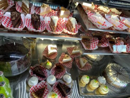 A display case filled with various cakes and desserts, including chocolate cake, cheesecake, and cupcakes, on checkered paper.