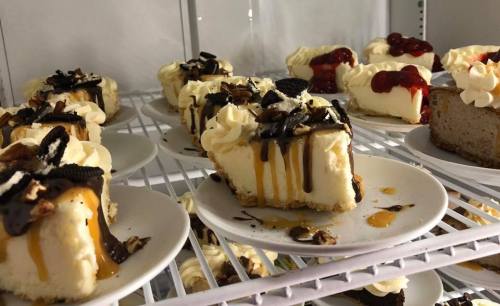 A display of various cheesecakes topped with whipped cream, chocolate, and fruit, on white plates in a refrigerator.