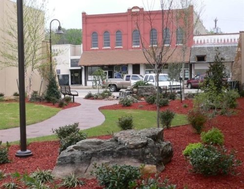 A landscaped park with a winding path, benches, and red mulch, featuring greenery and a historic building in the background.