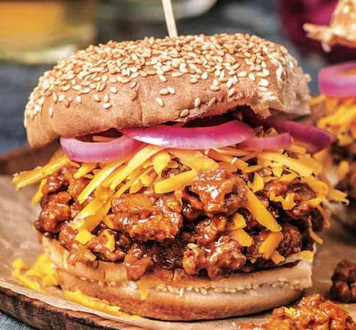 A close-up of a sesame seed bun filled with shredded cheese, onions, and a savory meat mixture on a wooden plate.