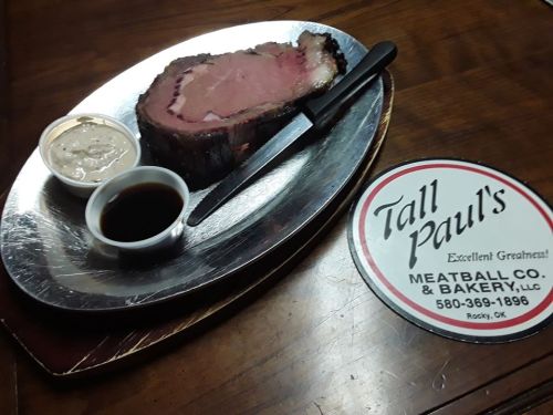 A plated slice of prime rib with dipping sauces, alongside a sign for Tall Paul's Meatball Co. & Bakery.