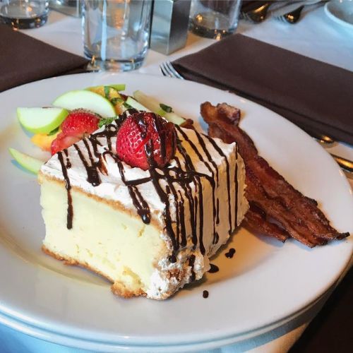 A slice of cheesecake topped with whipped cream, chocolate drizzle, a strawberry, and served with fruit and bacon.