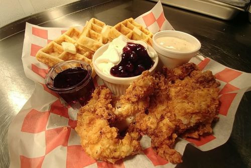 A plate featuring crispy fried chicken, waffles, whipped cream with cherries, and syrup in a checkered basket.