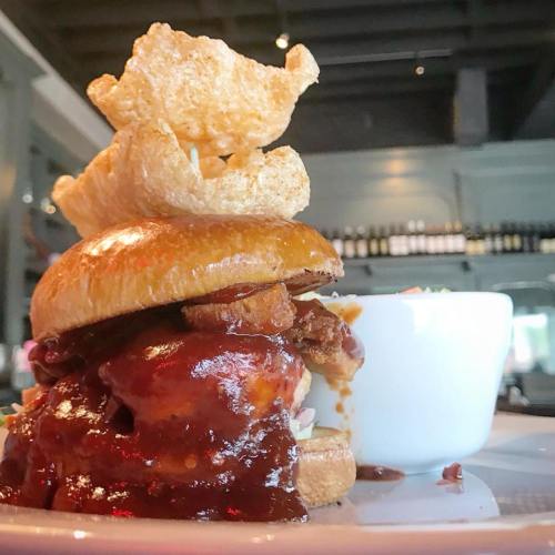 A stacked burger with crispy pork rinds on top, served with a side of coleslaw in a small bowl.