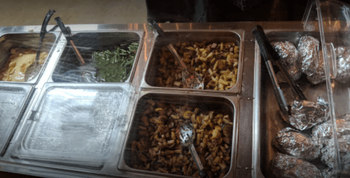 A food display with various dishes, including green beans, mixed vegetables, and foil-wrapped potatoes.